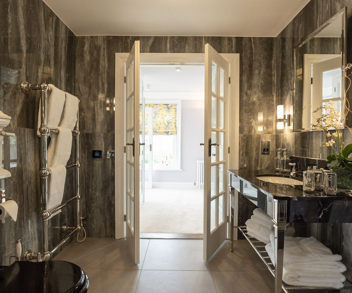A dramatic luxury bathroom with full dark marble wall cladding, chrome heated towel rail, black marble vanity unit and French doors - residential interior design by Atlantic Interiors