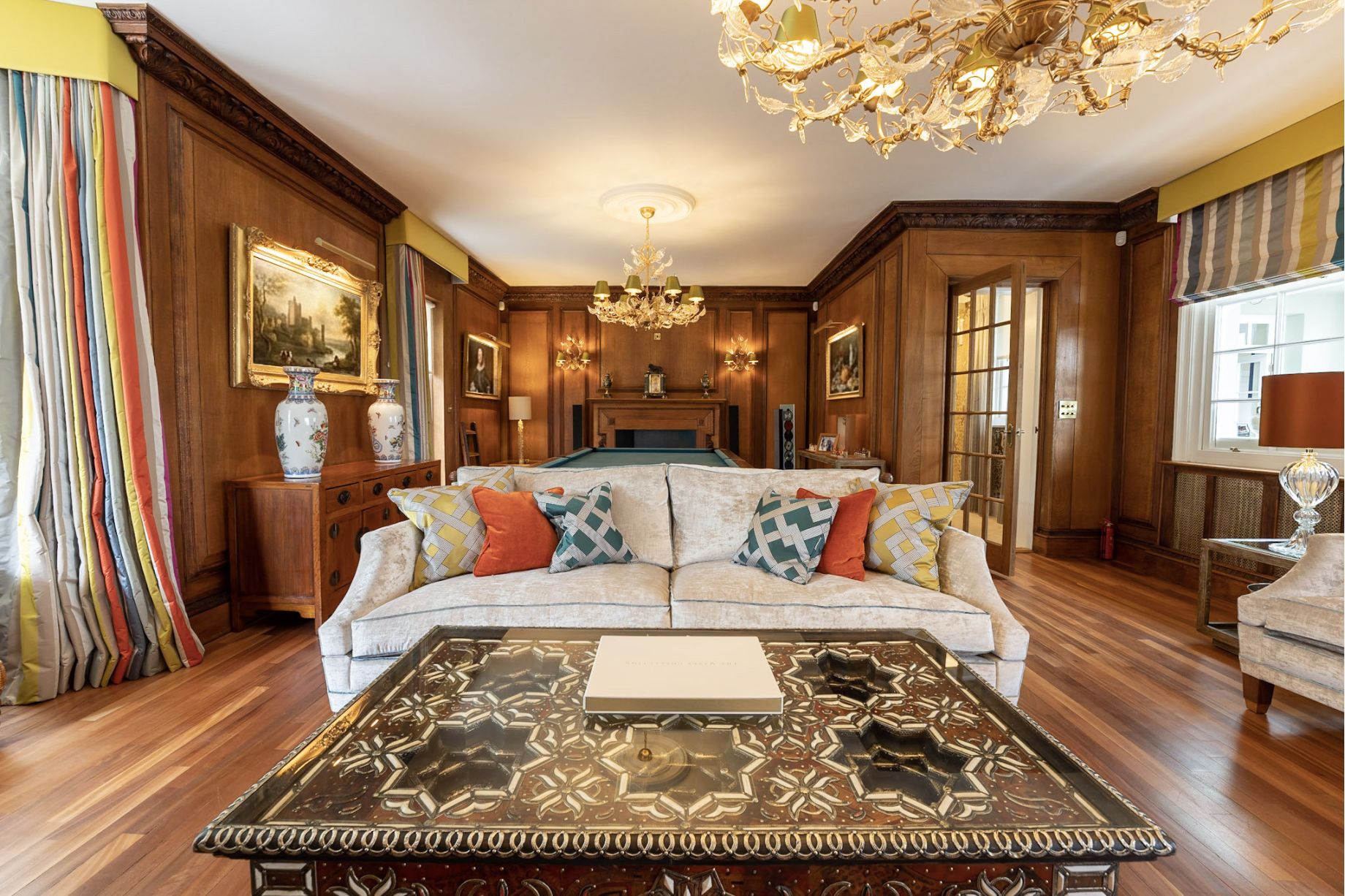 A grand drawing room with full dark oak wall panelling, ornate Murano chandelier, cream sofa with colourful cushions and a billiard table - residential interior design by Atlantic Interiors

