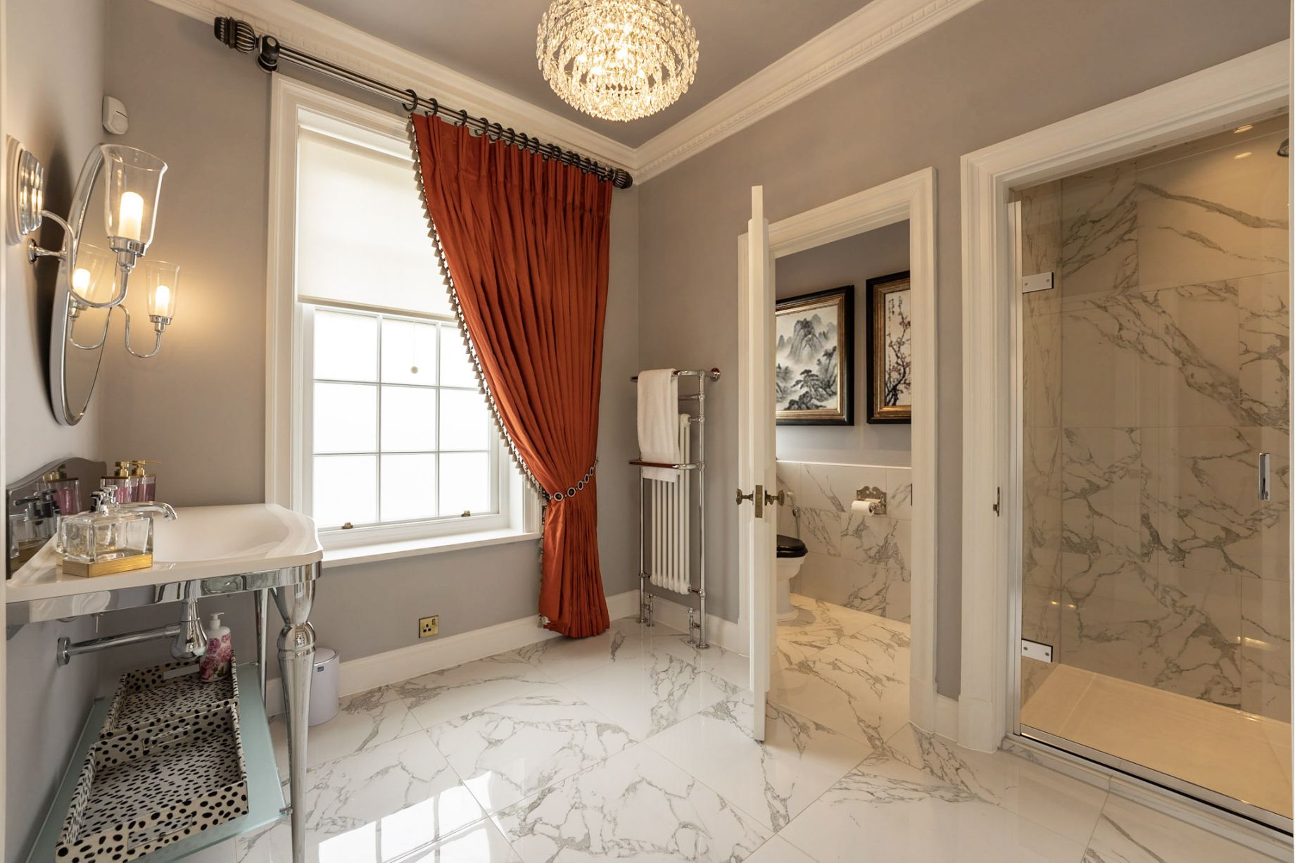 A luxury bathroom with bold orange silk curtains, a crystal chandelier, white marble floor tiles, a chrome console vanity and a view through to a marble shower room - residential interior design by Atlantic Interiors