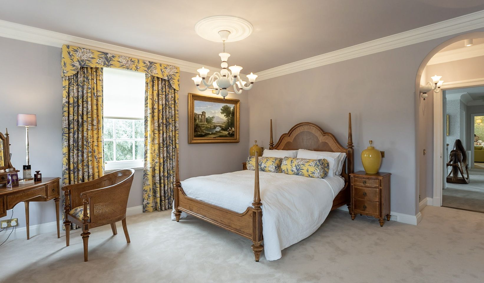 A luxury master bedroom with a four-poster walnut bed, yellow botanical curtains, a white Murano glass chandelier and yellow ceramic bedside lamps - residential interior design by Atlantic Interiors